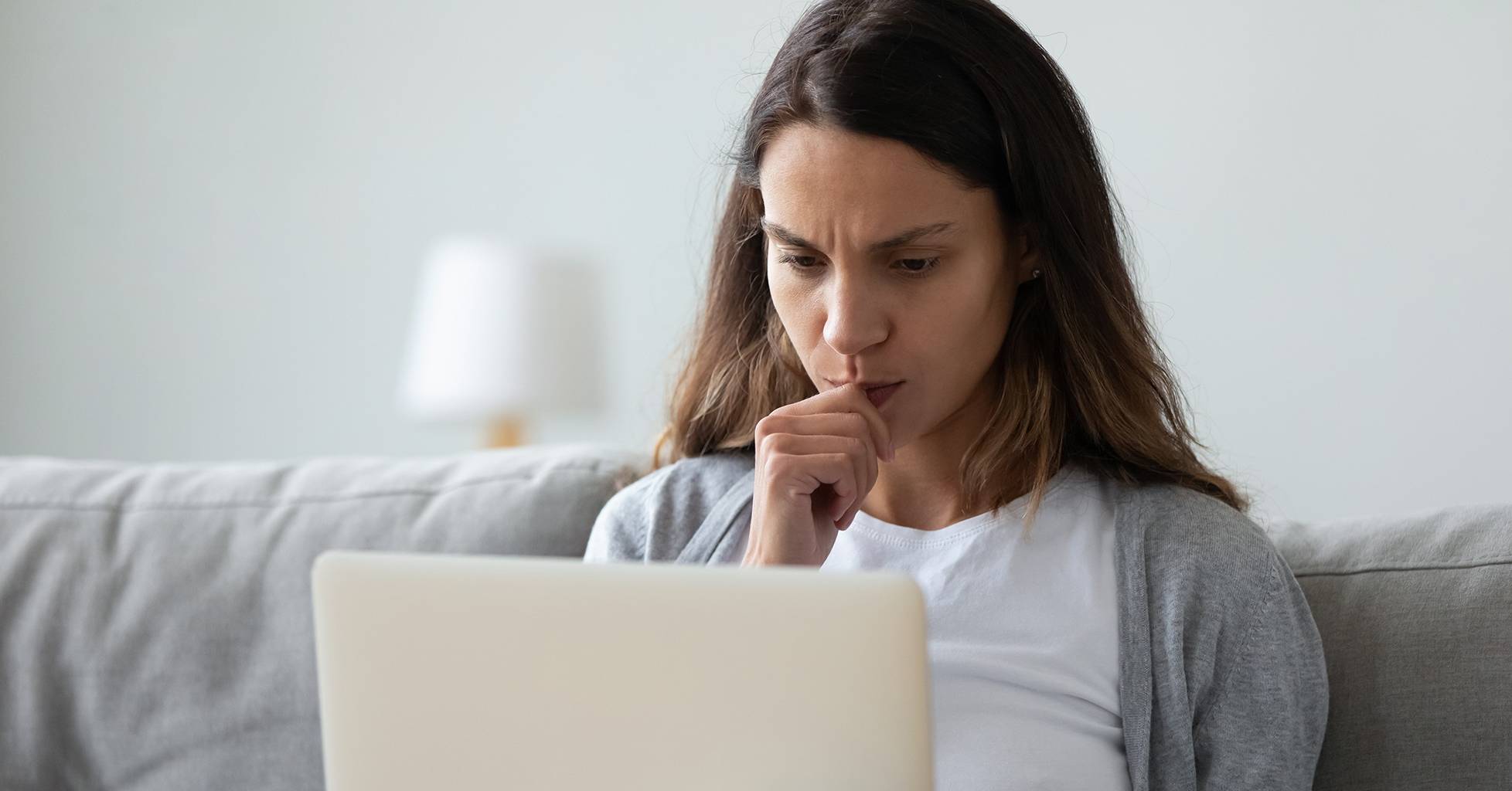 woman on couch facing laptop with closed hand on her lip and chin